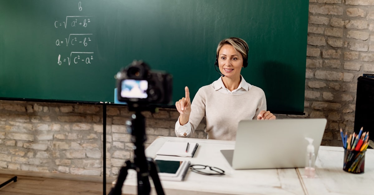 Educator wearing a headset records a classroom podcast at a desk with a laptop, camera, and chalkboard displaying math formulas.