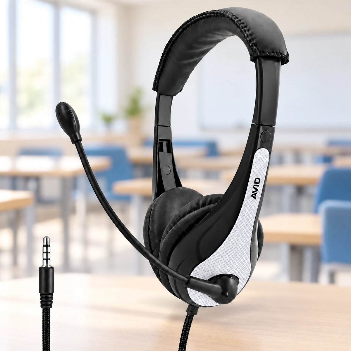 Black and white AVIO over-ear headset with an adjustable boom microphone, displayed on a wooden desk with a blurred classroom background.